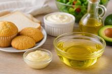 A bowl of golden cooking oil surrounded by baked goods, creamy spreads, salad, tomatoes, herbs, lemon and avocado, representing functional oils used in food products.