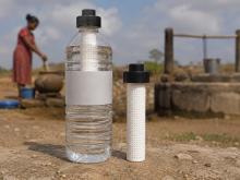 A clear plastic water bottle fitted with a white portable filter stands in the foreground beside a second filter cartridge, with a rural well and water collection scene in the background.