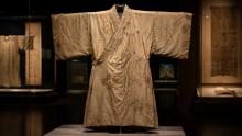 A pale embroidered silk robe displayed inside a glass museum case, illuminated against a dark exhibition background with other artifacts faintly visible.