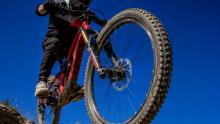 Close-up action photo of a mountain bike’s front tyre and fork leaping upward during a jump, ridden by a cyclist against a clear blue sky.