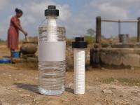 A clear plastic water bottle fitted with a white portable filter stands in the foreground beside a second filter cartridge, with a rural well and water collection scene in the background.