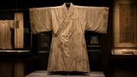 A pale embroidered silk robe displayed inside a glass museum case, illuminated against a dark exhibition background with other artifacts faintly visible.
