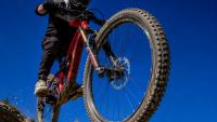 Close-up action photo of a mountain bike’s front tyre and fork leaping upward during a jump, ridden by a cyclist against a clear blue sky.